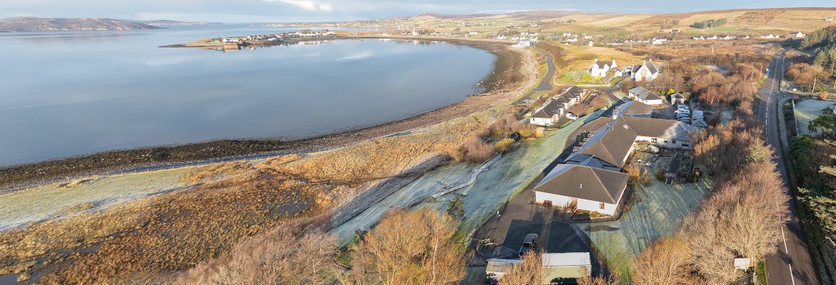 Aerial view of the Home, showing the sea bay on the left half and the buildings on the right half. A road borders the picture on the very right.